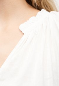 Κοντινό πλάνο ώμου με ανοιχτόχρωμο δέρμα και λευκό συγκεντρωμένο ύφασμα με φραμπαλά, που δείχνει την υφή και τις λεπτές πτυχώσεις του υφάσματος.