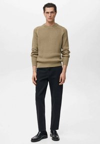 Beige gebreide trui met ribbelstructuur, ronde halslijn, gedragen met donkere, taps toelopende broeken en zwarte schoenen. Eenvoudig gestyled, minimale hardware.