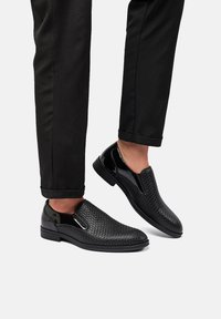 Scarpe slip-on nere con texture intrecciata, accenti lucidi e inserti elastici laterali. Abbinati a pantaloni neri a sigaretta.