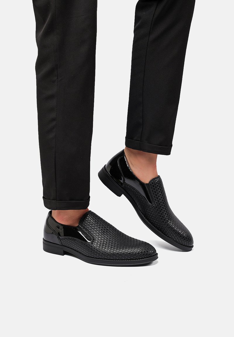 Scarpe slip-on nere con texture intrecciata, accenti lucidi e inserti elastici laterali. Abbinati a pantaloni neri a sigaretta.