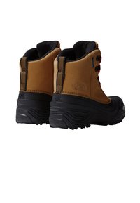 Cizme impermeabile maro din material sintetic, cu talpă neagră de cauciuc, guler căptușit și accente roșii la șireturi, având un logo pe călcâi și cusături detaliate.