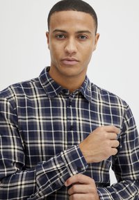 Cămașă în carouri navy și crem, confecționată dintr-un material cu guler butonat și mâneci lungi, având butoane negre și o textură netedă.