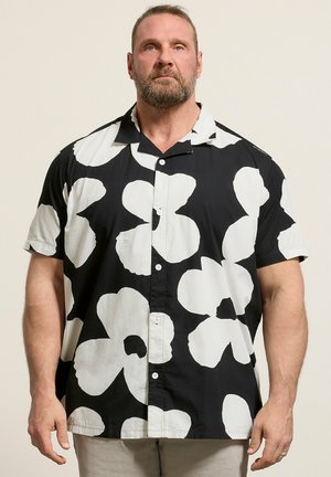 Mand med skæg iført en sort kortærmet skjorte med store hvide blomsterprint, stående mod en ensfarvet lys baggrund.