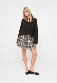 Dunkelgrüner, übergroßer Pullover mit V-Ausschnitt und weiten Ärmeln; karierten plissierten Rock in Grau und Braun; glänzende burgunderfarbene Ballerinas.