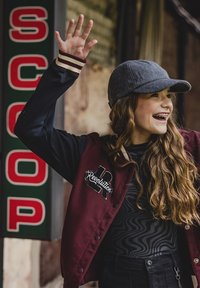 Bordeauxkleurige varsityjack met zwarte mouwen, voorzien van borduurwerk met de tekst "Revolution". Grijze pet, zwart top met golvend patroon, en een geheven hand.