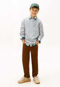 Maglione grigio a costa waffle sopra una camicia a quadri blu e verde, pantaloni di velluto a coste marroni e scarpe da ginnastica chiare; cappellino verde con logo.