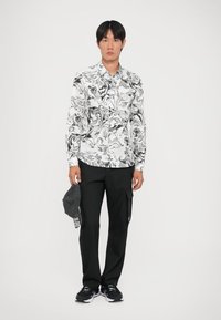 Camisa de botones blanca y negra con un patrón de remolinos, combinada con pantalones cargo negros y zapatillas negras. El modelo sostiene una gorra gris.