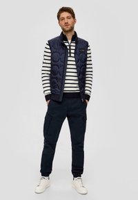 Navy quilted vest over a white and navy striped sweater, paired with dark cargo pants featuring pockets and elastic cuffs, completed with white sneakers.