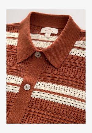 Dzianinowa koszulka polo w kolorze rdzy z białymi paskowanymi wycięciami. Posiada kołnierzyk na guziki i pojedyncze zapięcie na guzik.