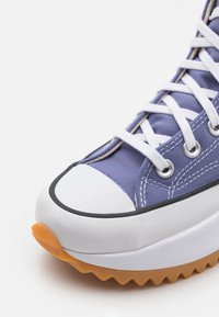 Lila canvas sneaker med vita snören, gummitåhättan och orange sula. Har silverfärgade öglor och kontrasterande sömnadsdetaljer.