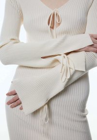 Cremfarbener, gerippter Strickpullover mit tiefem V-Ausschnitt, Kordelzugakzenten an Brust und Ärmeln sowie glockenförmigen Bündchen. Glatte Textur.