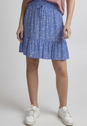 Jupe bleue à fleurs avec un ourlet volanté nouée à la taille, portée avec des baskets blanches et un top rose rentré.