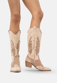 Jasno beżowe zamszowe kowbojki z ciemniejszymi dekoracyjnymi wzorami liści i bocznym zamkiem błyskawicznym, noszone na nagich nogach na białym tle.
