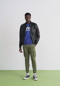 Black leather bomber jacket, blue sweatshirt, olive cargo pants, and black sneakers. Model stands on green flooring against a plain backdrop.