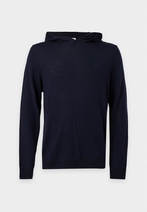 Homme debout portant un sweat à capuche noir, un pantalon beige et des mocassins en cuir noir sur un fond blanc uni.