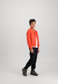 Orange bomber jacket with ribbed cuffs and collar, worn over a white shirt. Black cargo pants and brown athletic shoes with accents.