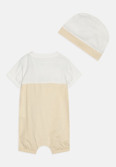 Mono de manga corta en algodón blanco y beige con un escote acanalado, y una gorra blanca con acentos beige a juego. Textura suave.