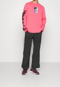 Chemise rose à manches longues avec un motif central, détails noirs sur les manches, associée à un pantalon noir ample et des baskets bordeaux.