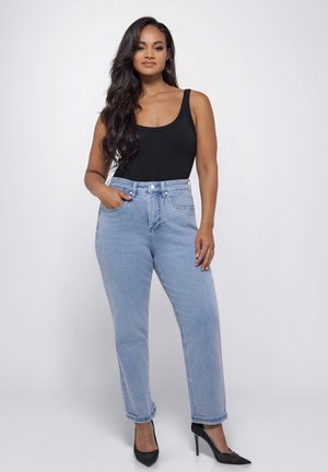Vrouw met lang donker haar, gekleed in een zwarte tanktop, lichtblauwe high-waisted jeans en zwarte hoge hakken, staand tegen een witte achtergrond.
