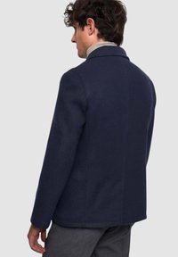 Cappotto blu navy in misto lana con vestibilità sartoriale, colletto con risvolto e spacco posteriore, mostrato di profilo con capelli ricci di media lunghezza.