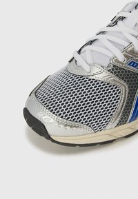Srebrna in črna športna čevlja z mrežastim zgornjim delom, črnimi gumijastimi podplati in modrimi detajli, ki imata zaobljen nos in ravne vezalke.