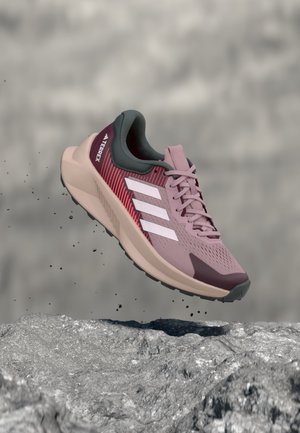 Chaussure de course rose et bordeaux avec des rayures blanches, en l'air au-dessus d'un terrain rocheux, petits débris tombant en dessous.