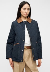 Μπουφάν καπιτονέ σε ναυτικό μπλε με καφέ γιακά και λεπτομέρειες, με κούμπωμα με κουμπιά μπροστά, πλαϊνές τσέπες και λεία υφή. Φορεμένο πάνω από λευκό τοπ και μπεζ παντελόνι.