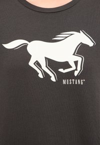 Černé tričko s bílou siluetou běžícího koně a slovem "MUSTANG" v bílém písmu, umístěné na tmavém pozadí.