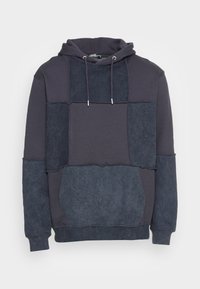 Felpa con cappuccio caratterizzata da un design a patchwork in diverse tonalità di blu scuro e navy, realizzata in morbido tessuto testurizzato con cordini regolabili.