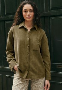 Camicia verde oliva, con bottoni, realizzata in un tessuto di cotone texturizzato. Presenta una tasca sul petto e una vestibilità rilassata con maniche lunghe.