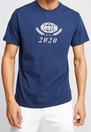 Homme portant un t-shirt Levi's bleu marine avec un logo blanc, des feuilles de laurier, les inscriptions « SF/CA » et « 2020 », associé à un pantalon blanc.