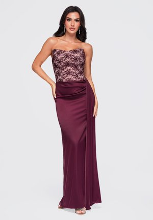 Femme debout dans une robe de soirée violette bustier avec un corsage texturé et un tissu drapé fluide, portant des talons hauts argentés et des boucles d'oreilles.
