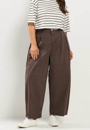 Femme portant un pantalon marron à jambes larges, des baskets blanches et une chemise blanche à manches courtes avec des rayures horizontales noires.
