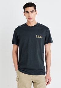 Tamsiai pilka medvilnės marškinėliai su apykakle ir trumpomis rankovėmis, su nusidėvėjusiu auksiniu "LEE" logotipu kairiajame krūtinės rajone.