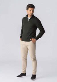 Mørkegrøn zip-up sweater, beige tætsiddende bukser, sorte sneakers. Slim fit, glat tekstur, afslappet design. Lys baggrund.