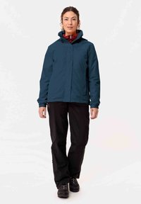 Giacca impermeabile di colore blu scuro con zip frontale, cappuccio e polsini elasticizzati, abbinata a pantaloni neri e stivali neri robusti.