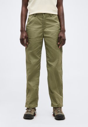 Άτομο φοράει χακί παντελόνι με ίσια γραμμή και παπούτσια πεζοπορίας, στέκεται μπροστά σε μια απλή ανοιχτή φωτεινή επιφάνεια.