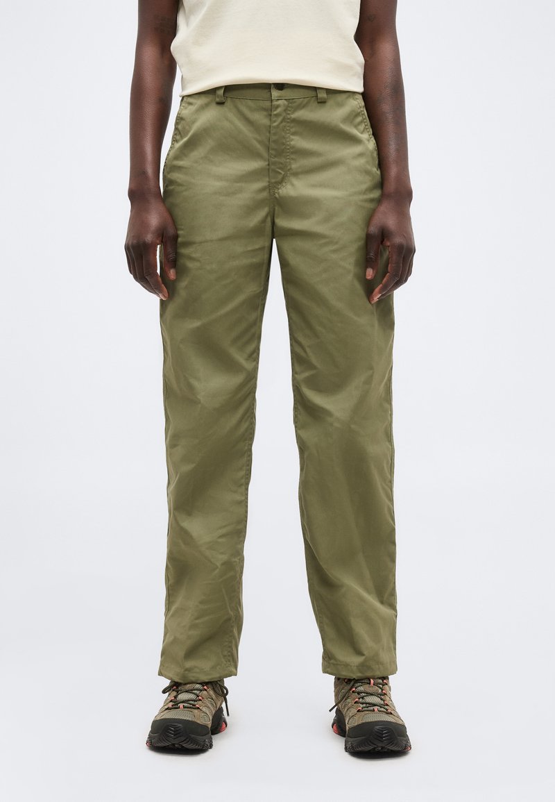 Asmuo, dėvintis alyvuogių žaliais tiesiais kelnais ir žygiavimo bateliais, stovintis prieš šviesų, vientisą foną.