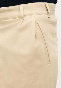 Pantalon beige avec passants de ceinture, poche latérale inclinée et coutures visibles, porté avec un haut noir rentré à la taille.