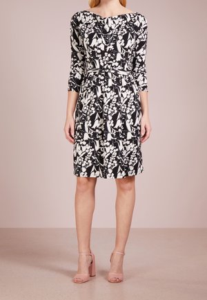 Vrouw die een knielange jurk draagt met zwarte en witte bloemenprint, driekwart mouwen en een dunne riem, gecombineerd met lichtroze sandalen met blokhakken.