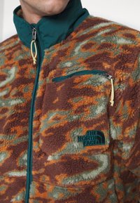 Homme portant une veste polaire marron, orange et verte à motifs, avec une poche poitrine zippée et le logo The North Face.