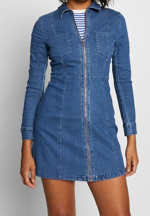 Frau trägt ein eng anliegendes blaues Jeanskleid mit langen Ärmeln, Reißverschluss vorne, Brusttaschen und Kragen über einem gestreiften Shirt.