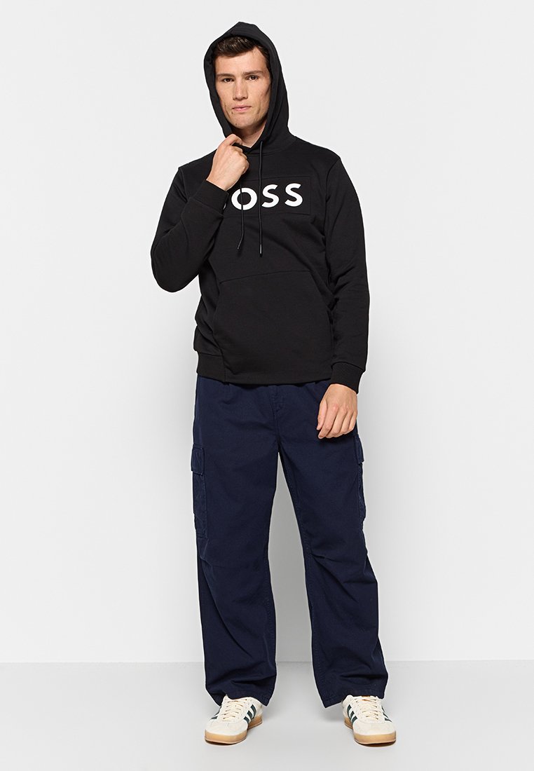 Homme portant un hoodie noir avec le logo "BOSS", un pantalon cargo bleu marine et des baskets blanches, se tenant devant un fond gris clair uni.
