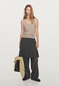 Žena v bezramínkovém béžovém topu, tmavé skladané sukni přes široké šedé kalhoty, držící béžový svetr a tmavě hnědou tašku.