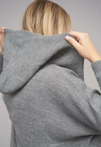 Grauer Strick-Hoodie mit einer großen Kapuze, aus strukturiertem Stoff und mit gerippten Bündchen, gehalten von zwei Händen von hinten.