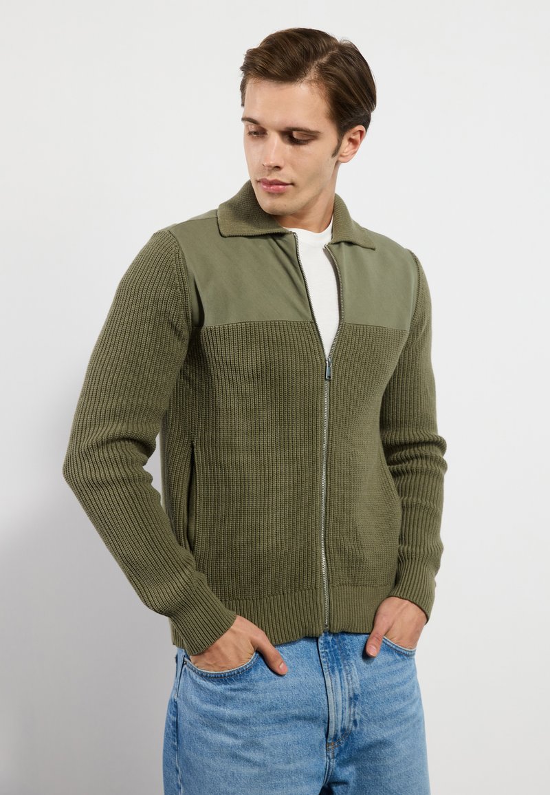 Sweatshirt zippé vert olive avec texture côtelée, section supérieure en tissu plat contrastant et deux poches latérales. Porté par-dessus une chemise blanche.