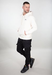 Sudadera crema con cordones y bolsillo central; combinada con pantalones cargo negros con múltiples bolsillos; deportivas negras con acentos grises.
