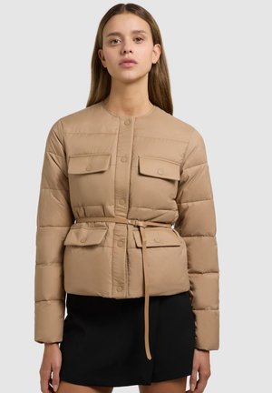 Kobieta ubrana w beżową pikowaną kurtkę z przednimi kieszeniami na klapę i cienkim paskiem, w połączeniu z czarną spódnicą, stojąca na jednolitym tle.