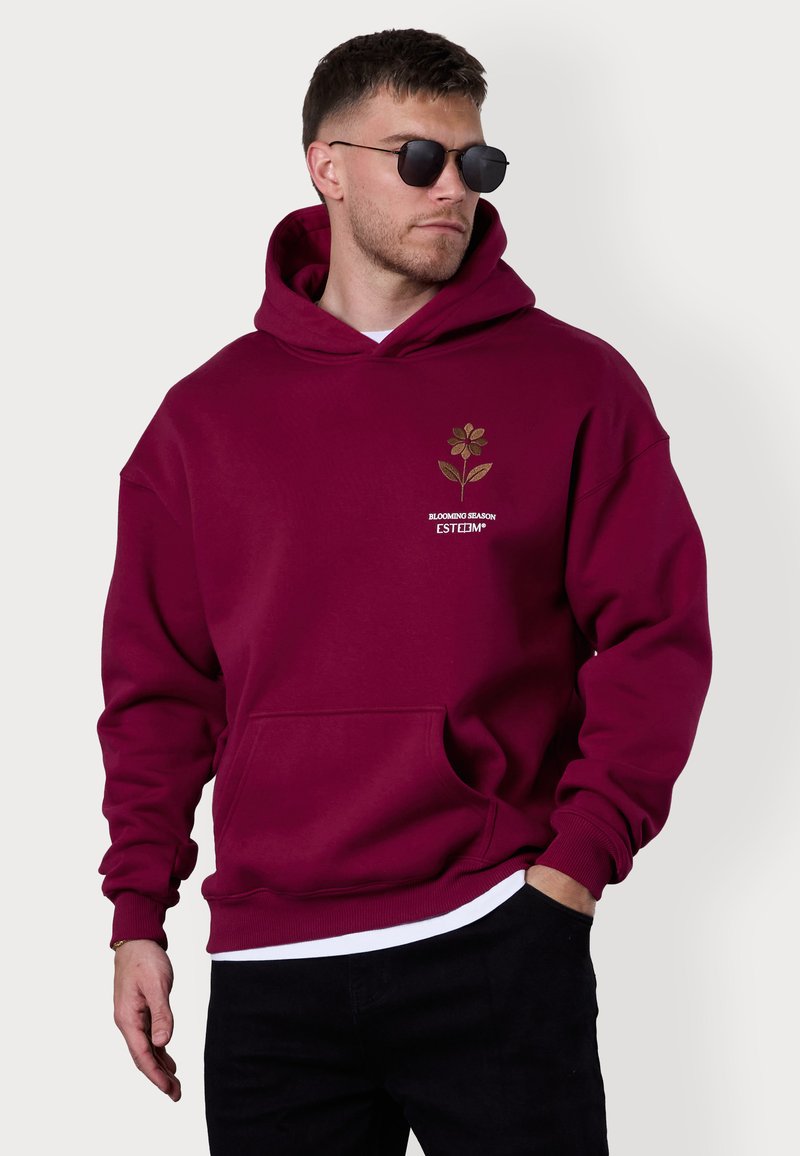 Bordeauxfarbener Hoodie aus weichem Material, mit einem Blumen-Graphic in Gold und weißem Text. Mit einer Fronttasche und gerippten Bündchen.