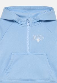 Gaiši zils kapucis ar priekšējo rāvējslēdzēju, kenguru kabatu un baltu "GAP" logotipu. Izgatavots no mīksta kokvilnas maisījuma auduma ar ribotiem rokturiem.